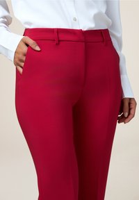 Pantalon rouge ajusté avec une texture lisse, poches latérales et une ceinture discrète ; accompagné d'une chemise blanche à manches longues.