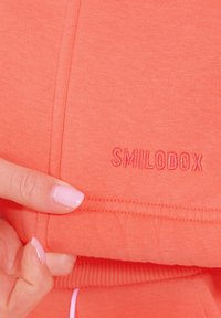 Tessuto atletico corallo con superficie testurizzata. Presenta il logo "SMILODOX" in un colore di filo coordinato, posizionato vicino all'orlo.