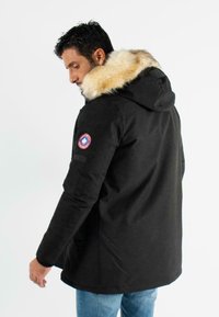 Parka noire avec une capuche en fourrure, arborant un patch logo sur la manche et une coupe légèrement ample. Texture lisse et matériau durable.