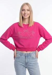 Jonge vrouw met steil blond haar die glimlacht, gekleed in een felroze cropped sweatshirt met de tekst "Happy Girls" en lichtblauwe spijkerbroek.