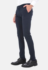 Pantaloni slim-fit navy con una texture liscia e tasche frontali, abbinati a scarpe nere stringate, presentati su uno sfondo neutro.