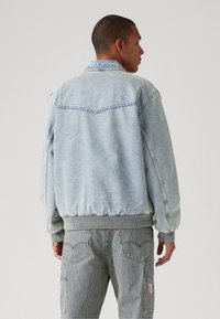 Veste en denim bleu clair avec un col, des poignets et un ourlet côtelés, et un détail au niveau du dos. Portée par-dessus un pantalon cargo rayé.