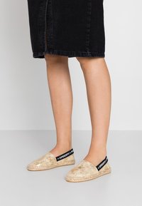 Espadrilles dorées avec une tige texturée, bande élastique noire étiquetée "SUPERDRY" et détail en jute sur la semelle. Portées avec une jupe en denim noire.