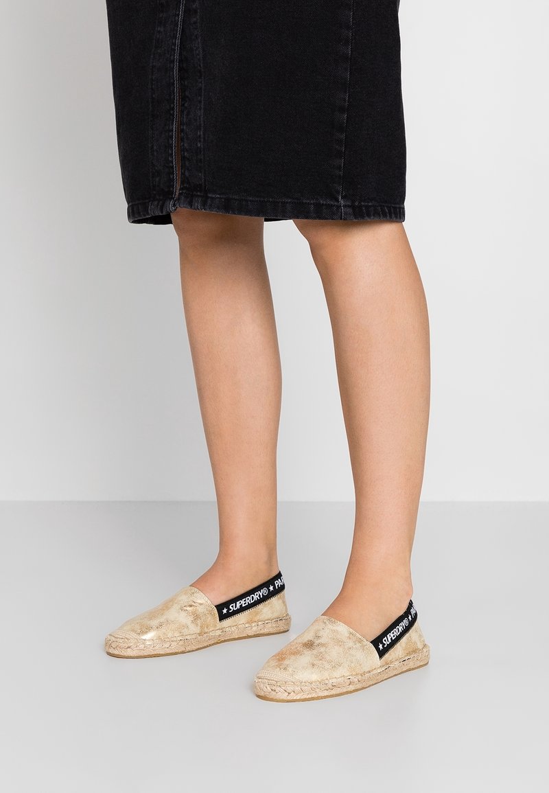Espadrilles dorées avec une tige texturée, bande élastique noire étiquetée "SUPERDRY" et détail en jute sur la semelle. Portées avec une jupe en denim noire.