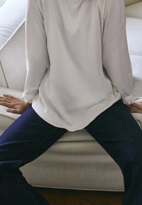 Haut blanc à manches longues avec des poignets cintrés, associé à un jean bleu foncé, assis sur un canapé de couleur claire avec une surface texturée.