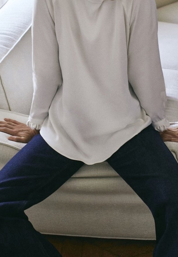 Haut blanc à manches longues avec des poignets cintrés, associé à un jean bleu foncé, assis sur un canapé de couleur claire avec une surface texturée.