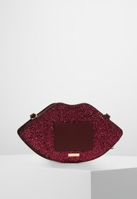 Metaliczna torebka w kolorze burgundowym z brokatem, w kształcie ust, z gładką, ciemnoczerwoną powierzchnią i małą złotą tabliczką z logo.
