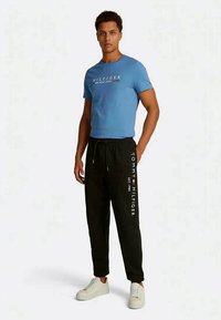 Homem vestindo uma t-shirt azul da Tommy Hilfiger e calças de moletom pretas com tênis brancos, de pé contra um fundo branco.