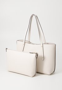 Nákladní taška z texturované kůže v off-white barvě s dlouhými uchy a odpovídajícím zipovým pouzdrem odpočívajícím vpředu na bílém pozadí.