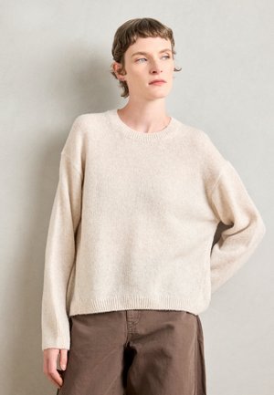 Lichtbeige gebreide trui met een ronde halslijn, verlaagde schouders en geribbelde zoom. Gekocht met bruine broek in een neutrale setting.