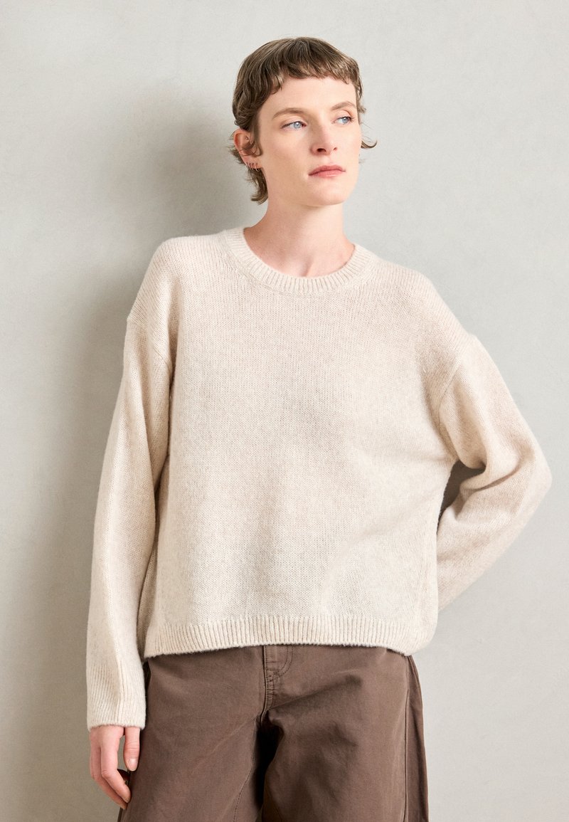 Lys beige strikkegenser med rund hals, senkede skuldre og ribbestrikket nederkant. Kombinert med brune bukser i en nøytral setting.