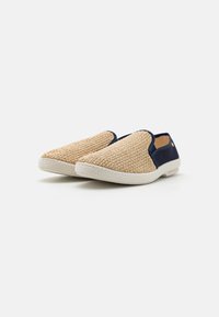 Chaussures slip-on avec une tige tissée beige, des accents en tissu bleu marine et une semelle en caoutchouc blanche. Présente un design texturé de forme arrondie.