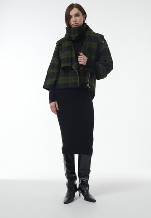 Chaqueta corta de cuadros verdes y negros con cuello oversized, combinada con un suéter negro de punto y una falda lápiz negra larga, acompañada de botas altas hasta la rodilla.