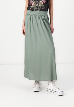 Femme portant une longue jupe vert clair, un t-shirt noir à motif floral et des baskets montantes blanches sur fond blanc.