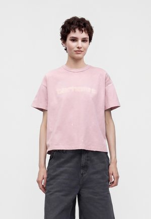 Jeune personne aux cheveux courts et bouclés portant un t-shirt ample rose Carhartt et un pantalon sombre taille haute sur un fond uni.