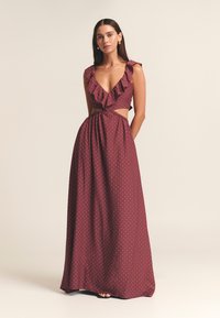 Abito maxi bordeaux con pois bianchi, scollatura arricciata, lati intagliati e dettaglio con lacci sul retro. Realizzato in tessuto leggero, dal design fluido.