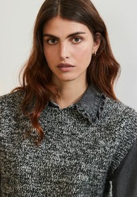 Strikket sweater i sort og hvid, med en ribbet tekstur og en grå denimkrave med bølgede kanter. Nærbillede af en persons overkrop.