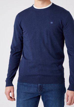 Námořnický modrý pletený svetr s dlouhými rukávy a kulatým výstřihem. Má strukturovanou tkaninu a malé logo na levé hrudi.