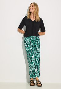 Musta lyhythihainen paita, jossa on V-pääntie ja tekstuuriset yksityiskohdat, yhdistettyinä vihreisiin kuvioituihin housuihin, joissa on geometrisiä muotoja ja mustia yksityiskohtia.