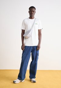 Camiseta blanca de algodón con logo verde, combinada con pantalones cargo de mezclilla azules y holgados con bolsillos laterales, zapatillas grises y una bolsa cruzada gris.