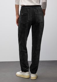 Persona che indossa pantaloni di velluto a coste neri con vita elastica e tasche posteriori, abbinati a sneaker bianche su un pavimento grigio.