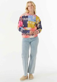 Bunter Sweater mit einer Mischung aus geometrischen und floralen Mustern in Pink, Blau, Gelb und Schwarz, kombiniert mit hellblauen Jeans und beigen Schuhen.