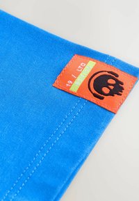 Sinine kangast plaastriga, millel on oranž etiket musta koljuga logo ja tekst "19 / OMAKAPANETA VERSIOON." Äärte ümber on nähtav õmblus.
