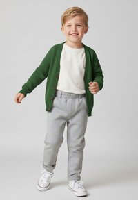 Zielony sweter rozpinany na guziki w brązowym kolorze, biały t-shirt, szare spodnie dresowe oraz białe trampki z czerwonymi akcentami. Prosty, casualowy strój.