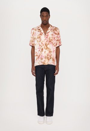 Chemise à manches courtes avec un motif tie-dye rose, marron et crème ; associée avec un jean bleu foncé et des baskets blanches.