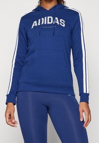 Žena v modré mikině značky Adidas s bílými pruhy na rukávech a odpovídajících modrých legínách na jednolitém pozadí.