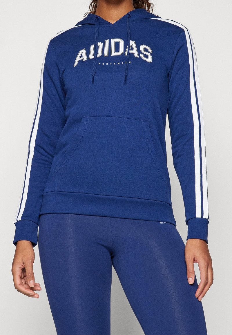 Žena v modré mikině značky Adidas s bílými pruhy na rukávech a odpovídajících modrých legínách na jednolitém pozadí.