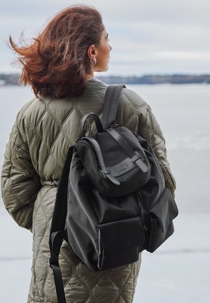 Schwarzer Stoff-Rucksack mit Klappenverschluss, verstellbaren Trägern und Fronttaschen. Die Tasche hat eine glatte Textur und minimale Markenkennzeichnung.