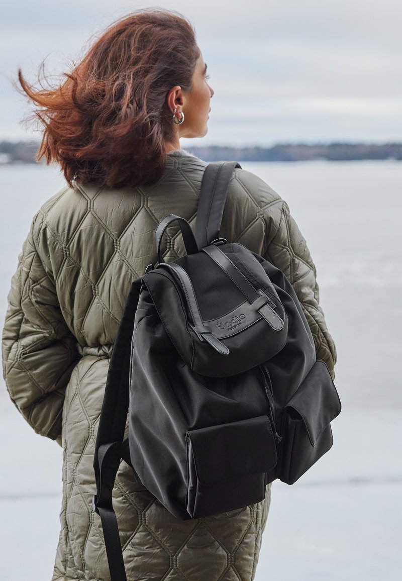 Svart tyg ryggsäck med lockstängning, justerbara remmar och framfickor. Väskan har en slät yta och minimal märkning.