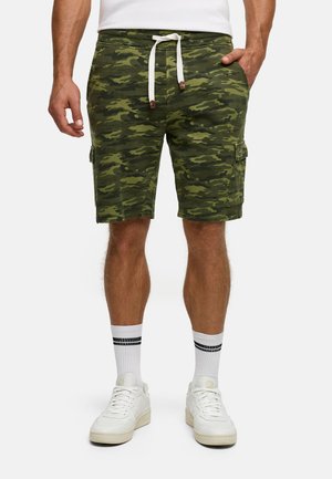 Mann trägt grüne Tarn-Cargo-Shorts mit weißem Kordelzug, weiße Socken mit schwarzen Streifen und weiße Sneaker und steht mit den Händen in den Taschen.
