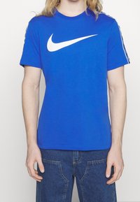 Henkilö, jolla on päälläsininen lyhythihainen paita, jossa on valkoinen Nike-merkki rintamuksessa, ja tummansiniset farkut, taustanaan yksinkertainen valkoinen tausta.