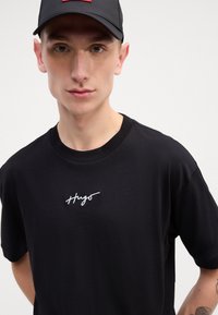 Czarna bawełniana koszulka z okrągłym dekoltem, z białym haftowanym logo "Hugo" na klatce piersiowej. Czapka z siateczkowym tyłem i płaskim daszkiem.