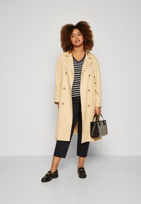 Trench beige con botones de doble botonadura, llevado sobre una camiseta de rayas en blanco y negro, combinado con pantalones cropped azul marino y un bolso a rayas.