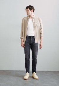 Camisa de botón color beige claro sobre una camiseta sin mangas blanca, combinada con jeans oscuros y zapatillas crema. Tejido suave, corte relajado y diseño sencillo.