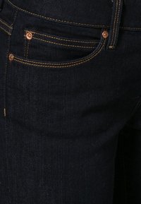 Jeans in denim blu scuro con accessori in rame, cuciture visibili e una tasca frontale. Il tessuto ha una trama liscia.