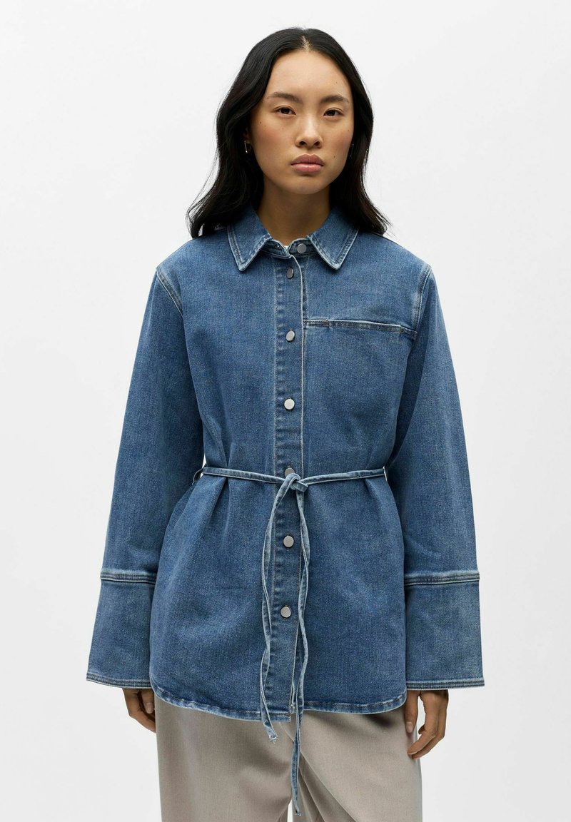 Femme portant une veste en jean à manches longues boutonnée avec ceinture à nouer, debout devant un fond gris clair uni.