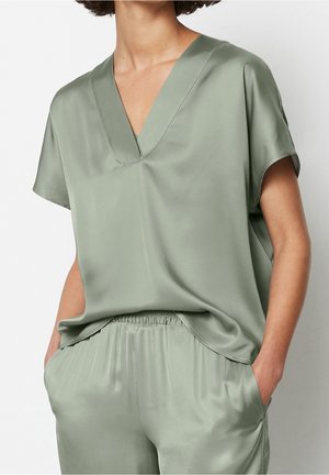Personne portant un haut ample à manches courtes vert clair avec col en V et un pantalon assorti à taille élastique, mains dans les poches, sur fond uni.