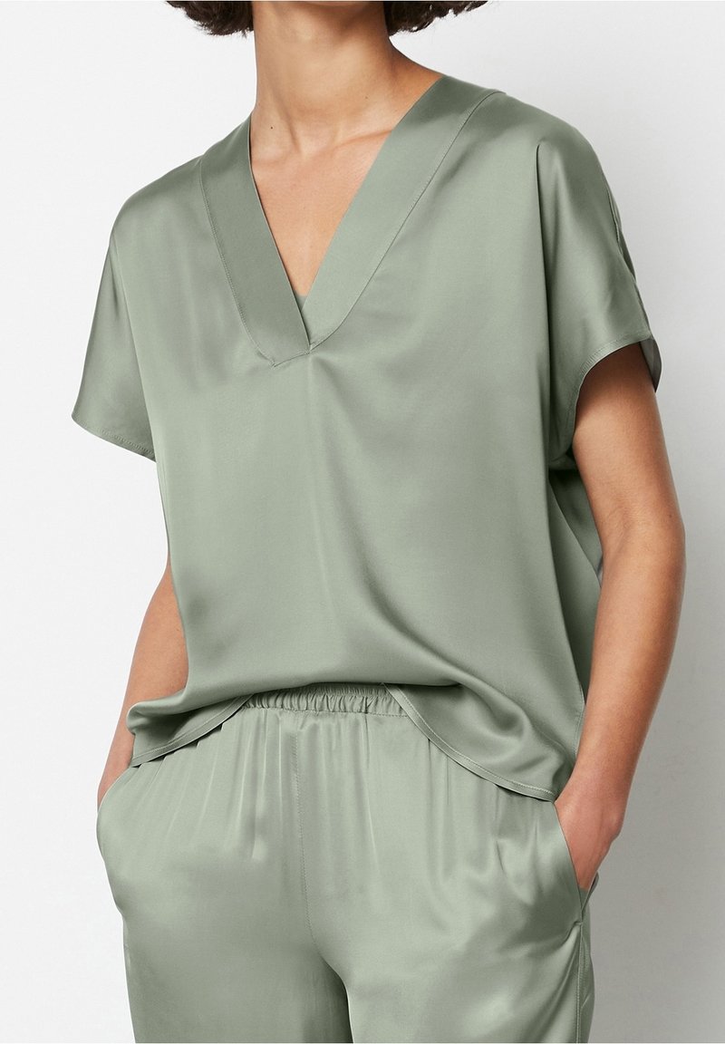 Personne portant un haut ample à manches courtes vert clair avec col en V et un pantalon assorti à taille élastique, mains dans les poches, sur fond uni.