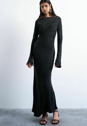 Mujer de pie con un vestido largo negro ajustado con sutiles rayas de cebra, mangas acampanadas y tacones abiertos negros, frente a un fondo sencillo.