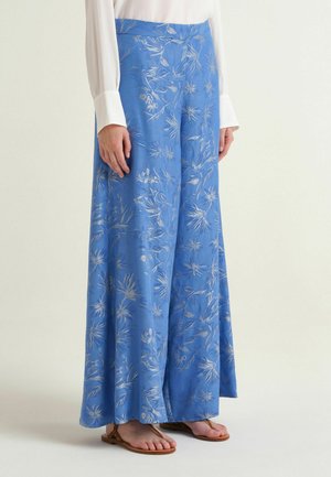 Pantaloni blu a gamba larga con stampa floreale bianca indossati con camicia bianca a maniche lunghe e sandali bassi marroni su sfondo semplice.