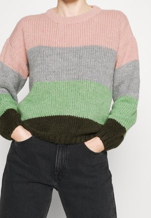 Person trägt einen gestrickten Pullover mit horizontalen Streifen in Pink, Grau, Hellgrün und Dunkelgrün, Hände in den Taschen einer schwarzen Hose.