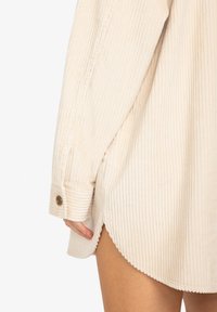 Gros plan sur une personne portant une chemise en velours côtelé beige avec des poignets boutonnés, montrant la couture latérale et l'ourlet festonné au-dessus de la cuisse nue.