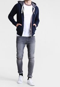 Sudadera con cremallera azul marino y forro gris, cordones blancos; camiseta blanca, vaqueros ajustados gris con roturas; zapatillas blancas con detalles negros.