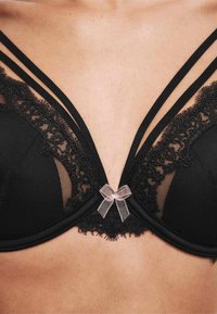 Czarny koronkowy stanik z przezroczystymi panelami, z paskami, falbankami i małą różową kokardką na środku.