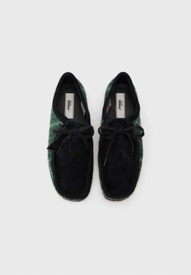 Black suede shoes with green woven accents, featuring a rounded shape, top laces, and stitched detailing along the edges.