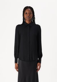 Black long-sleeve shirt with a button-down collar, featuring vertical pleats and a smooth texture. Worn with a matching dark skirt.